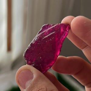 May include: A translucent, deep magenta-colored piece of raw glass with an irregular, jagged shape and a textured, uneven surface. The material catches the light, displaying varying shades of dark pink and red throughout its crystalline structure.