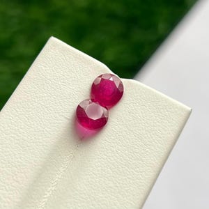 Può includere: Due rubini rotondi sfaccettati di un colore rosso intenso e vibrante. Le pietre preziose sono posizionate su un cartoncino espositivo testurizzato color bianco sporco. Ogni rubino presenta un taglio brillante con molteplici sfaccettature che riflettono la luce.