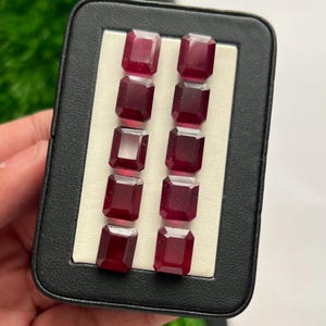 Puede incluir: Una colección de diez piedras preciosas de color rojo intenso, con corte esmeralda, dispuestas en dos filas verticales de cinco dentro de un estuche expositor rectangular negro. Cada piedra presenta superficies facetadas que reflejan la luz.