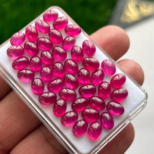 May include: A collection of numerous small, oval-shaped, vibrant pinkish-red gemstones arranged in a grid pattern on a white foam insert within a clear rectangular plastic display case. Each gemstone features a smooth, polished cabochon surface.