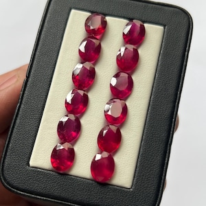Puede incluir: Una colección de doce rubíes rojos de corte ovalado y facetado, dispuestos en dos filas verticales dentro de un estuche rectangular negro con forro interior blanco. Cada piedra preciosa exhibe un tono carmesí profundo y brillante.