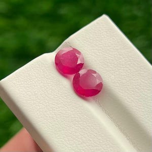 Op de afbeelding: Twee ronde, gefacetteerde, levendig roze-rode edelstenen op een wit gestructureerd oppervlak. Elke steen heeft een ronde slijpvorm met meerdere facetten die het licht reflecteren, wat een diepe, verzadigde kleur en een gladde afwerking toont.