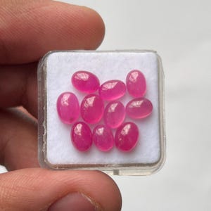 Könnte beinhalten: Eine Ansammlung von zehn kleinen, ovalen, leuchtend pinkfarbenen Edelsteinen, die in einem quadratischen weißen Schaumstoffeinsatz in einer durchsichtigen Kunststoffbox angeordnet sind. Jeder Stein hat eine glatte, polierte Cabochon-Oberfläche.