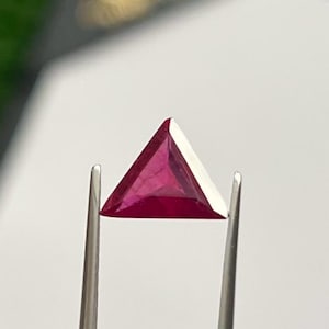 Peut inclure: Un rubis triangulaire et facetté, tenu par une pince argentée. Le rubis est d'un rouge profond et saturé avec une facette blanche sur un côté. La gemme est posée sur un fond blanc flou.
