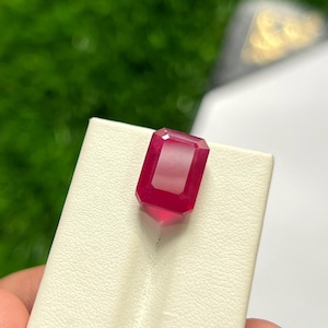 Peut inclure: Une pierre précieuse d'un rouge profond et vibrant, taillée en forme d'émeraude rectangulaire avec des bords facettés. La pierre présente une surface claire et polie, posée sur un support de présentation blanc cassé texturé.