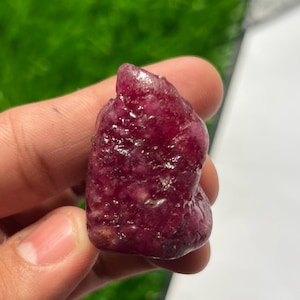 Puede incluir: Una piedra preciosa de rubí en bruto y sin pulir, de color rojo magenta intenso con una superficie texturizada e irregular. La piedra tiene una forma ligeramente irregular y redondeada con facetas cristalinas naturales.