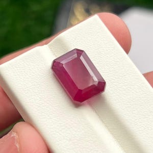 May include: A faceted, rectangular ruby gemstone with a deep red hue. The gem is displayed on a white surface, showcasing its clarity and brilliance. The cut of the stone highlights its facets, reflecting light and creating a captivating visual effect.