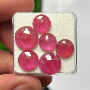 May include: Five round, faceted pink gemstones arranged in a clear plastic display case with a white foam insert. Each stone features a geometric cut with multiple triangular facets that reflect light, showcasing a vibrant, consistent pink color.