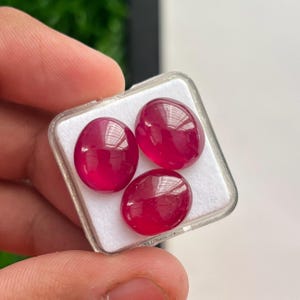 Könnte beinhalten: Drei ovale, tief magentarote Cabochon-Edelsteine mit einer glatten, polierten Oberfläche. Die Steine sind in einer dreieckigen Formation in einer kleinen, quadratischen, transparenten Kunststoff-Präsentationsbox mit weißem Schaumstoffboden angeordnet.