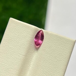 Op de afbeelding: Een marquise-geslepen edelsteen met een levendige roze tint en een gefacetteerd oppervlak. De steen is geplaatst op een getextureerde, gebroken witte displaystandaard, die de langwerpige, puntige vorm en interne lichtreflecties toont.