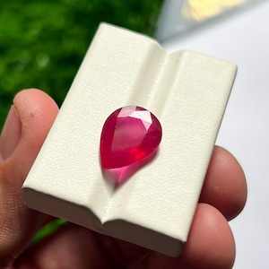 May include: A pear-shaped, faceted gemstone with a vibrant, deep red colour. The stone is set against a white, rectangular display card that features a central vertical groove, providing a clean contrast to the gemstone's brilliant red hue.