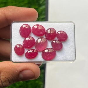 Könnte beinhalten: Eine Sammlung von neun polierten, oval geformten, tief rosa-roten Edelsteinen, die auf einem weißen Schaumstoffeinsatz in einer durchsichtigen, rechteckigen Kunststoff-Präsentationsbox angeordnet sind. Die Steine haben eine glatte, reflektierende Oberfläche.