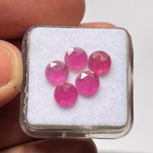 Peut inclure: Cinq pierres précieuses rondes, facettées, rose vif sont présentées dans un étui en plastique transparent avec un intérieur blanc. Les gemmes ont une taille brillante, reflétant la lumière. L'étui est tenu par des doigts.