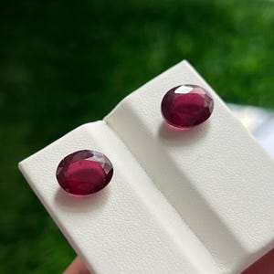 Op de afbeelding: Twee ovaal geslepen, dieprode edelstenen met gefacetteerde oppervlakken. De stenen worden gepresenteerd op een witte, getextureerde en schuine standaard. Elke edelsteen vertoont een rijke, verzadigde karmozijnrode tint met lichtreflecties.