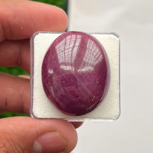 May include: A polished, oval-shaped cabochon gemstone with a deep magenta and purple hue. The stone features a smooth, reflective surface with visible internal inclusions and natural veining, securely set within a square white display case.