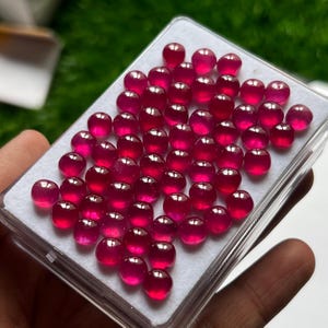 Op de afbeelding: Een rechthoekige doorzichtige plastic displaydoos met een verzameling van talrijke kleine, ronde, gepolijste, dieproze-rode edelstenen. De stenen zijn in een rasterpatroon gerangschikt op een witte schuimrubberen inzet in de doos.