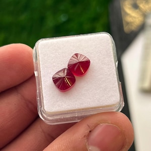 Può includere: Due piccole pietre preziose rosse di forma quadrata, sfaccettate con una superficie superiore a piramide. Le pietre sono esposte all'interno di un contenitore quadrato in plastica trasparente con un inserto in schiuma bianca.