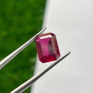 May include: A rectangular, faceted gemstone with a deep, vibrant red color held by a pair of silver-toned metal tweezers. The stone features a clear, polished surface with sharp edges and a distinct geometric cut that reflects light across its facets.