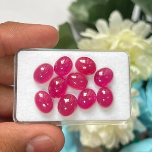 Könnte beinhalten: Eine Sammlung von neun ovalen, im Cabochon-Schliff geschliffenen, leuchtend pinkfarbenen Edelsteinen, die in einer rechteckigen, durchsichtigen Kunststoffschatulle mit weißer Schaumstoffeinlage angeordnet sind. Jeder Stein hat eine glatte Oberfläche.