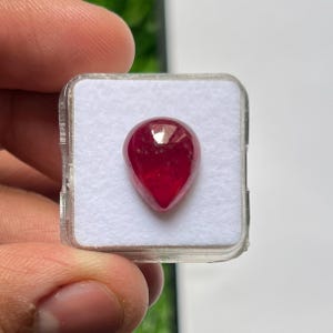 Op de afbeelding: Een peervormige, dieprode edelsteen met een glad, gepolijst oppervlak. De steen is stevig gecentreerd in een vierkante, doorzichtige plastic displaydoos met een witte, getextureerde schuiminzet om de edelsteen op zijn plaats te houden.