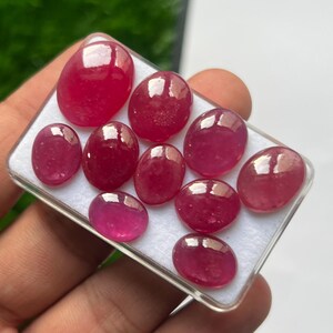 May include: A collection of ten polished, oval-shaped, deep pinkish-red gemstones arranged in a clear plastic display box with a white foam insert. Each stone features a smooth, rounded cabochon cut with a reflective surface and varying sizes.