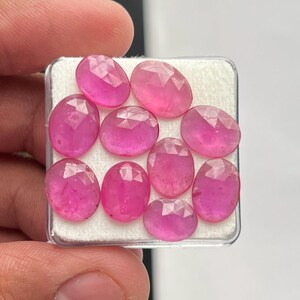 Op de afbeelding: Een verzameling van negen ovaalvormige, gefacetteerde roze edelstenen gerangschikt in een vierkante witte displaydoos. Elke steen heeft een gepolijst oppervlak met geometrische facetten die het licht reflecteren en een levendige kleur tonen.