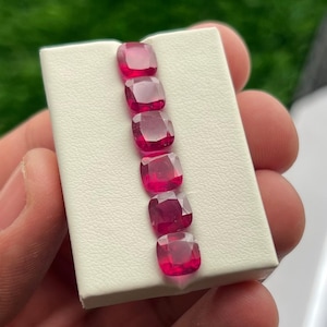 Könnte beinhalten: Sechs kissenförmig geschliffene Rubine, von tiefroter Farbe, werden auf einer weißen Oberfläche präsentiert. Die Facetten der Edelsteine sind sichtbar und reflektieren das Licht. Die Rubine sind vertikal angeordnet und heben ihre Form und Farbe hervor.