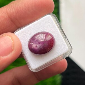 以下が含まれることがあります： 滑らかで光沢のある表面を持つ、深いマゼンタ色の楕円形のカボションカットの宝石。この宝石は、しっかりと固定するための白いフォームインサートを備えた、小さな正方形の透明なプラスチック製のディスプレイボックスの中に安全に収められています。