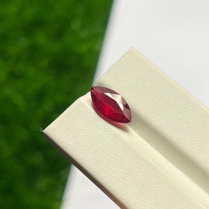 May include: A marquise-cut ruby gemstone with a deep red color and faceted surface. The stone features a pointed oval shape with reflective surfaces that catch the light, displaying a vibrant crimson hue throughout its transparent structure.