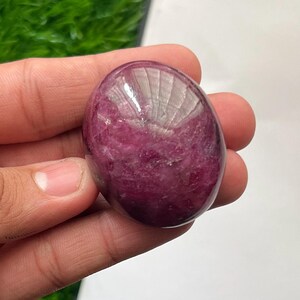 Op de afbeelding: Een gepolijste, ovaalvormige edelsteen met een diepe magenta en paarse tint. De steen heeft een glad, reflecterend oppervlak met zichtbare interne kristallijne texturen en natuurlijke variaties in kleurintensiteit door de hele vorm.