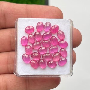 Könnte beinhalten: Eine Sammlung ovalförmiger, rosa Edelsteine, präsentiert in einer transparenten Kunststoffbox mit weißem Innenraum. Die polierten Edelsteine haben ein glänzendes Aussehen und variieren leicht in der Farbgebung.