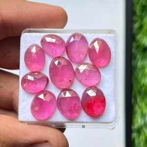 May include: Nine oval-cut, pink gemstones are presented in a clear plastic case. The gems have a faceted surface, catching the light to create a sparkling effect. The pink hues vary, with some stones displaying a deeper red.