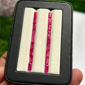Può includere: Due file verticali di gemme vibranti color rosa intenso con taglio baguette sono esposte in una custodia rettangolare nera con un inserto in velluto bianco. Ogni fila contiene sette pietre rettangolari disposte in linea retta.