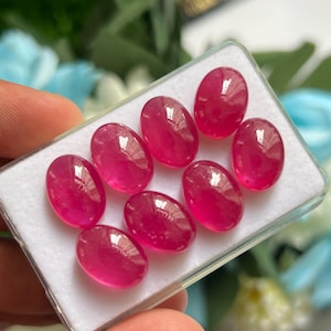 Könnte beinhalten: Eine Sammlung von sieben ovalen, durchscheinenden, leuchtend pinkfarbenen Edelsteinen, die in einer durchsichtigen Kunststoff-Präsentationsbox mit weißer Schaumstoffeinlage angeordnet sind. Jeder Stein hat einen glatten, polierten Cabochon-Schliff.