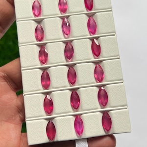 May include: A collection of sixteen marquise-cut pink gemstones arranged in a white display tray with horizontal slots. Each gemstone features a faceted surface and a vibrant, translucent pink hue, uniformly positioned in four rows of four stones.