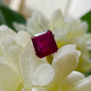 May include: A deep magenta, rectangular-cut gemstone set in a simple metal prong setting. The stone features a faceted surface that reflects light, showcasing a rich, saturated colour. The gemstone is positioned centrally, displaying clear edges.