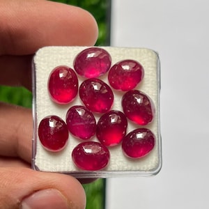 May include: A collection of nine oval-shaped, deep pinkish-red gemstones arranged on a white foam insert within a clear plastic display box. Each stone features a smooth, polished cabochon surface that reflects light, showcasing a vibrant color.