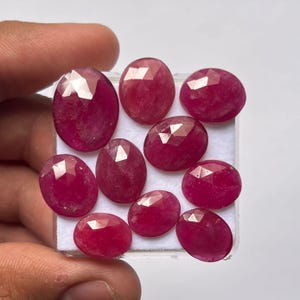 Puede incluir: Colección de nueve rubíes ovalados y facetados. Las piedras preciosas son de un color rojo intenso y rico, presentadas sobre una superficie blanca. Las facetas capturan la luz, creando un efecto brillante. Estos rubíes son ideales para la fabricación de joyas.