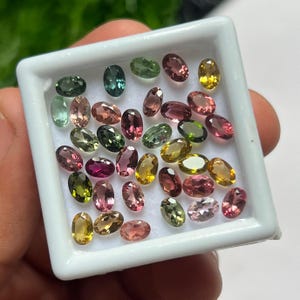 May include: A collection of small, oval-cut loose gemstones in a white square display box. The stones feature a variety of colors including shades of pink, green, yellow, and brown, all with a faceted surface that reflects light across the display.