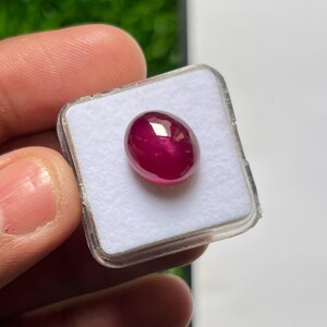Può includere: Una gemma ovale di colore rosso magenta intenso con un taglio cabochon liscio e lucido. La pietra è posizionata su un inserto in schiuma bianca all'interno di una scatola espositiva in plastica trasparente, con una superficie riflettente.
