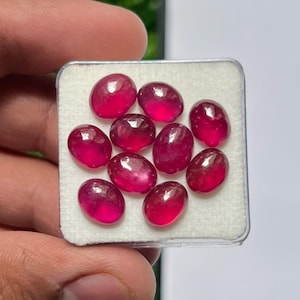 Könnte beinhalten: Eine Ansammlung von neun ovalen, im Cabochon-Schliff geschliffenen Rubinen mit tiefroter Farbe und glatten, polierten Oberflächen. Die Edelsteine sind in einer quadratischen, durchsichtigen Kunststoff-Präsentationsbox mit weißem Schaumstoff angeordnet.