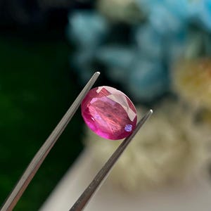 May include: A vibrant, oval-cut pink gemstone held by a pair of silver-toned metal tweezers. The stone features a faceted surface that reflects light, showcasing a deep magenta hue with internal clarity and distinct geometric patterns across its top.