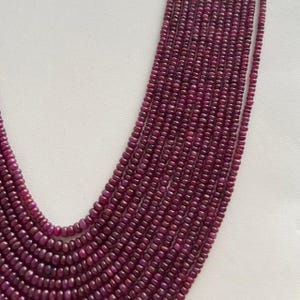 Pode incluir: Um colar de várias voltas composto por pequenas contas redondas de cor vermelho escuro. As contas são enfiadas juntas, criando um efeito de cascata. O colar é exibido sobre um fundo branco liso.