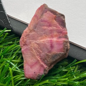 Pode incluir: Uma gema natural em bruto, com uma mistura de tons rosa e castanho-avermelhado. A pedra tem uma superfície texturizada e uma forma irregular, com áreas de rosa mais intenso e secções mais claras, quase translúcidas. A gema é exibida sobre um leito de relva artificial verde.