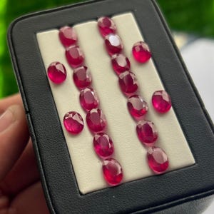 May include: A selection of oval-cut, deep red rubies presented in a black jewellery box. The gemstones are arranged on a white surface, highlighting their polished facets and rich colour. The box is held by a hand, suggesting a presentation or sale.