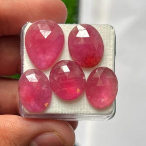 Puede incluir: Cinco piedras preciosas rosas facetadas dispuestas en una caja de presentación de plástico transparente con un inserto de espuma blanca. La colección incluye una piedra en forma de pera y cuatro piedras ovaladas, todas con facetas.