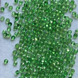 May include: A large collection of small, round, faceted green gemstones. Each individual stone features a brilliant cut with multiple reflective surfaces, creating a sparkling appearance across the entire group of loose, vibrant emerald-coloured gems.