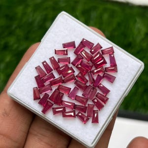 Puede incluir: Una colección de pequeñas piedras preciosas facetadas de color rojo rosado intenso, talladas en forma de baguette cónica. Estas piedras sueltas y translúcidas están dispuestas en un grupo sobre una base de espuma blanca en un estuche.