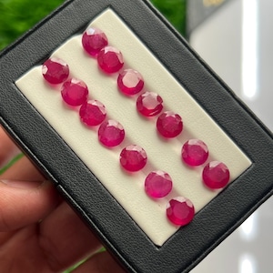 Puede incluir: Una colección de catorce piedras preciosas redondas, facetadas y de un vibrante color rojo rosado, dispuestas en dos filas paralelas dentro de una caja de presentación negra con un inserto de espuma blanca. Cada gema tiene un corte circular.