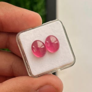 May include: Two oval-shaped, translucent pink gemstones with a smooth, polished cabochon cut. The stones are displayed side-by-side on a white foam insert inside a small, square, clear plastic display box with a hinged lid.